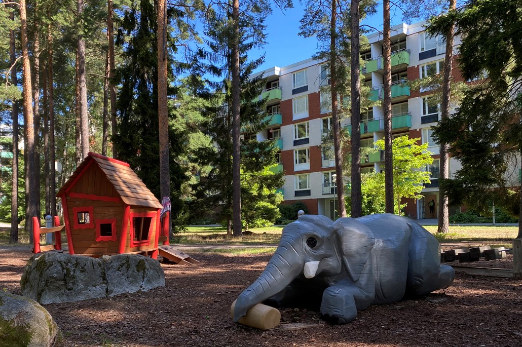 Lekpark med en grå elefant som ligger på mage och en röd lekstuga i en dunge med tallar framför ett flerbostadshus i vitt, grönt och brunt