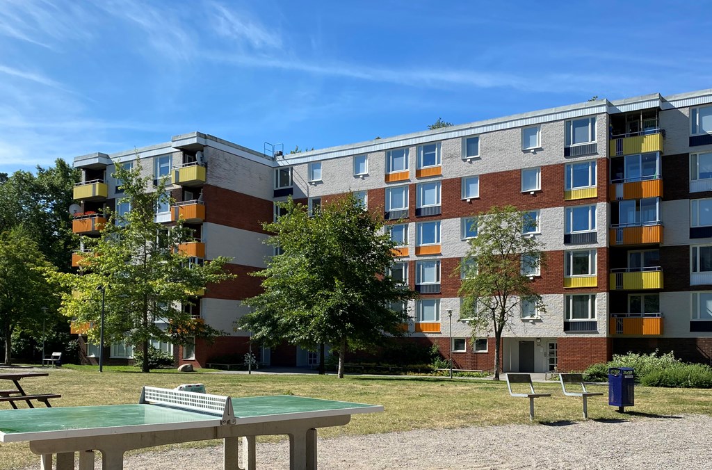 Pingisbord i förgrunden med en gräsmatta och träd framför ett flerbostadshus i flera våningar i vitt, brunt och orange