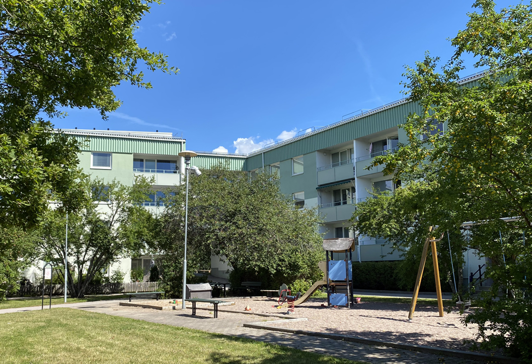 Ljusgrönt flerbostadshus i vinkel omslutande en gård med gräsmatta, träd och lekpark