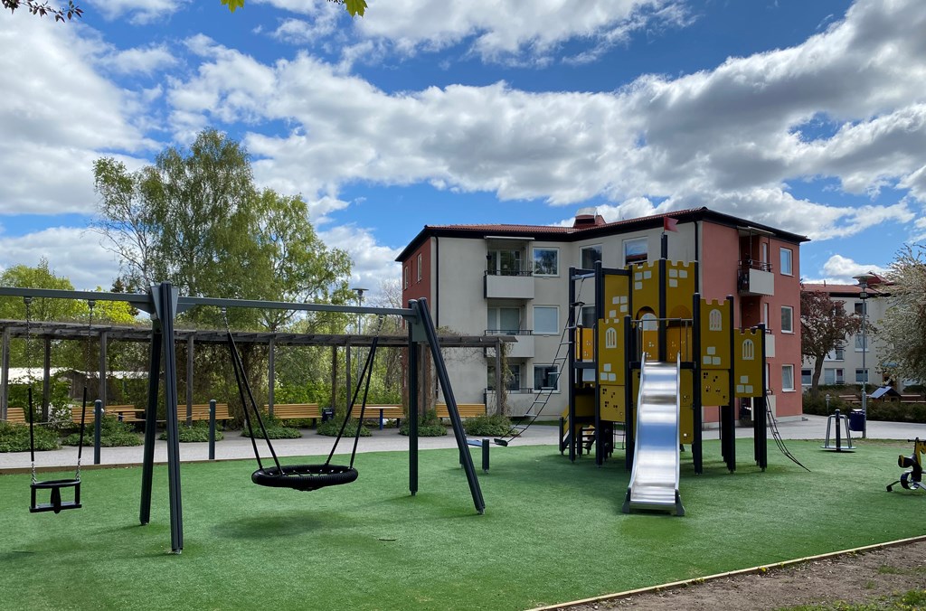 Lekpark med gungor, klätterställning med rutschkana framför ett flerbostadshus i vitt och rosa