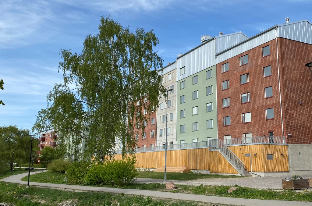 Flerbostadshus i rött, grönt och grått med ett staket invid en gångväg med träd