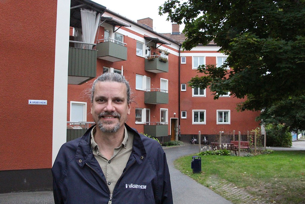 Man i blå jacka framför flerbostadshus i rött och vitt med gröna balkonger samt en gräsmatta och tr