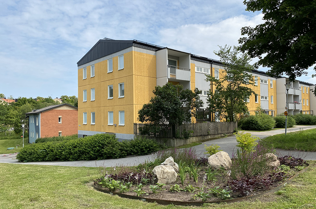 Gult flerbostadshus med gräsmatta och rabatt i förgrunden