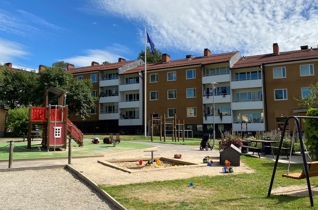 Gult flerbostadshus i tre våningar med vita balkonger och i förgrunden en lekplats med sandlåda och rutschkana