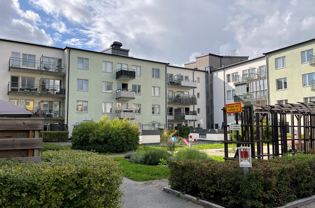 Grönt och vitt flerbostadshus i vinkel kring en grönskande innergård med buskar och lekplats.