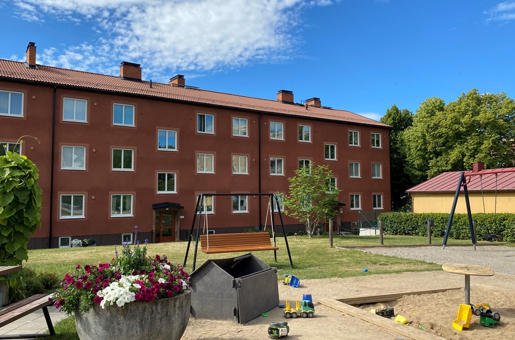 Brunfärgat trevånings flerbostadsshus med bruna balkonger med en gräsmatta och en lekplats, blommor och bänkar i förgrunden