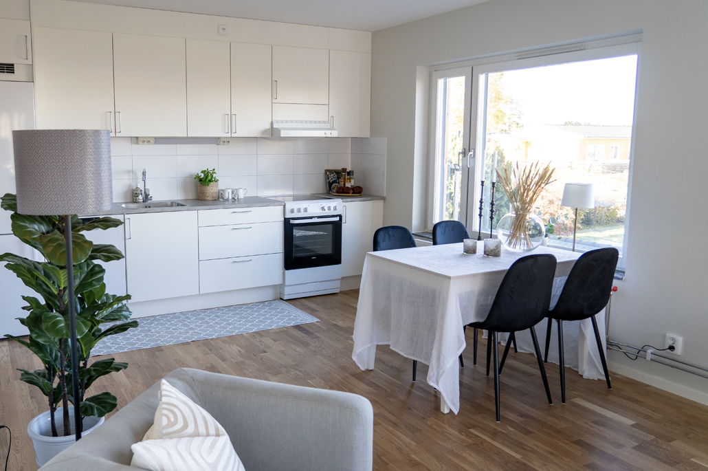 Köksvägg med vita skåpluckor, ekparkett, matbord med vit duk och svarta stolar framför ett fönster och en grå fåtölj och grön växt i förgrunden.