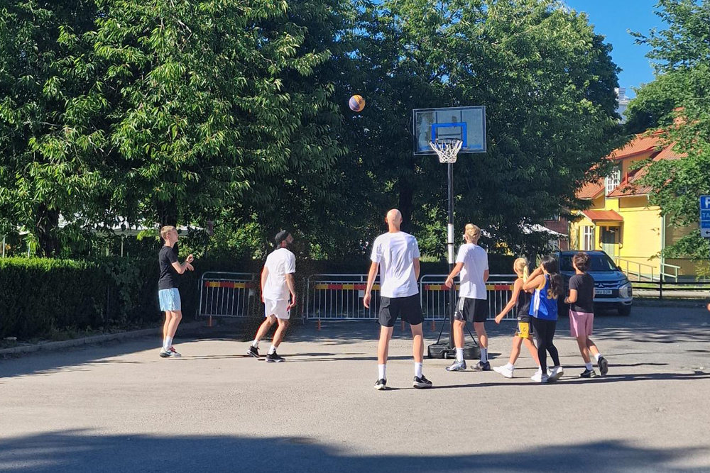 Ungdomar som spelar basket med ett gult trähus och gröna träd i bakgrunden.