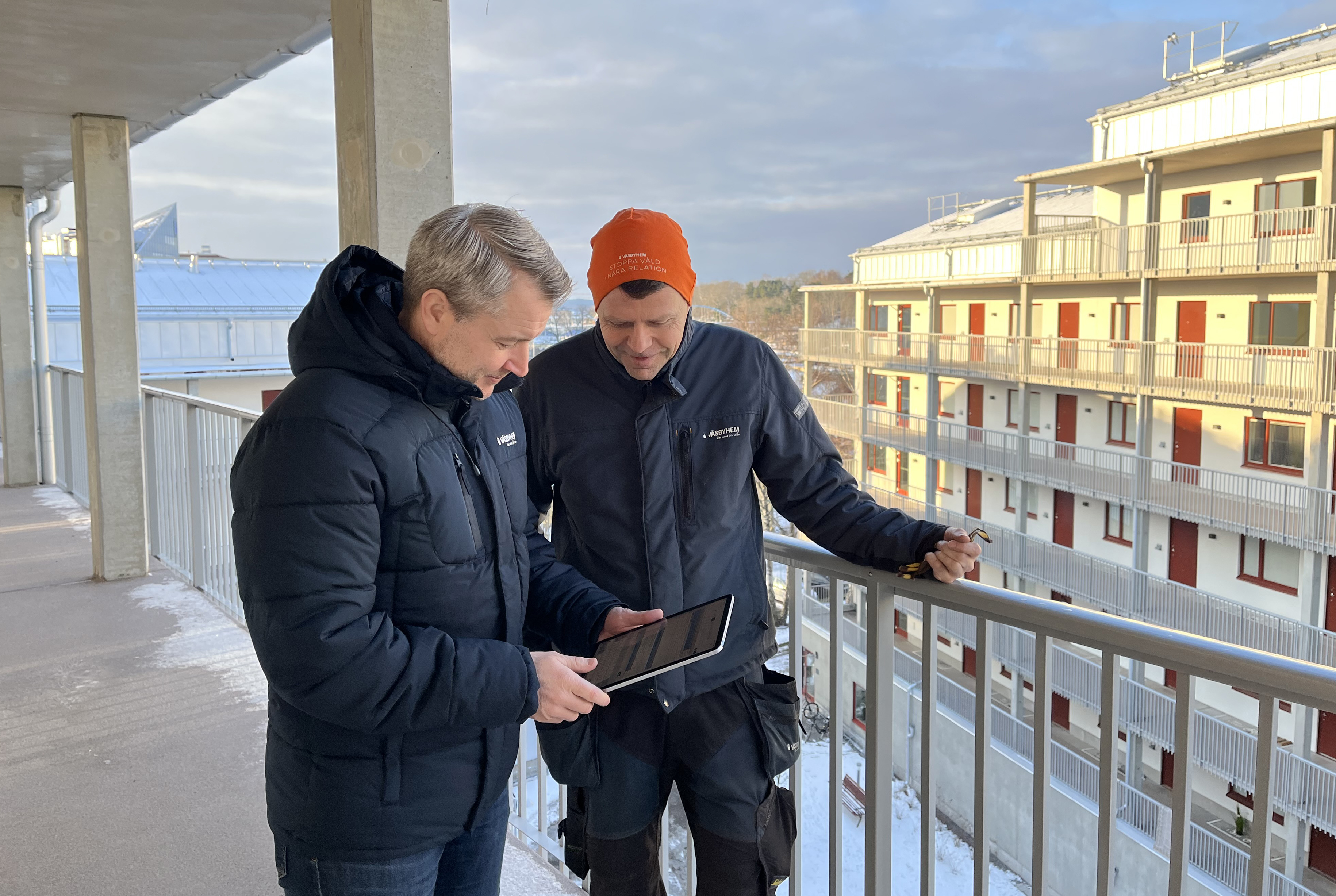 Två män står i arbetskläder på en loftgång med ett annat flerbostadshus i bakgrunden.