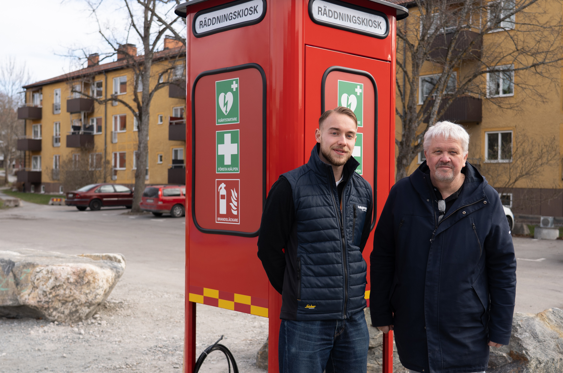 Två män står framför en röd räddningskiosk som ser ut om en gammal telefonkiosk. 