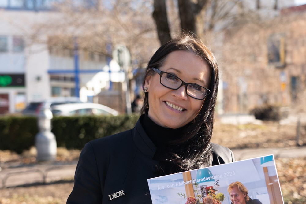 Kvinna i mörka kläder och glasögon stående utomhus med en års- och hållbarhetsrapport i händerna.