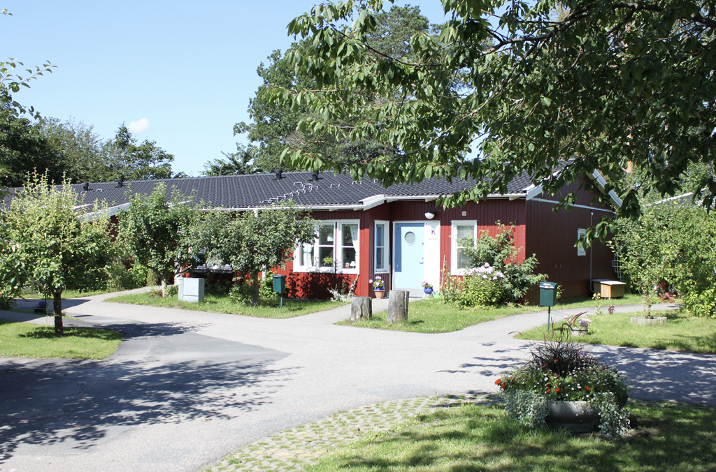 Rött enplansradhus i rödmålat trä med gångvägar, träd och blomlåda i förgrunden