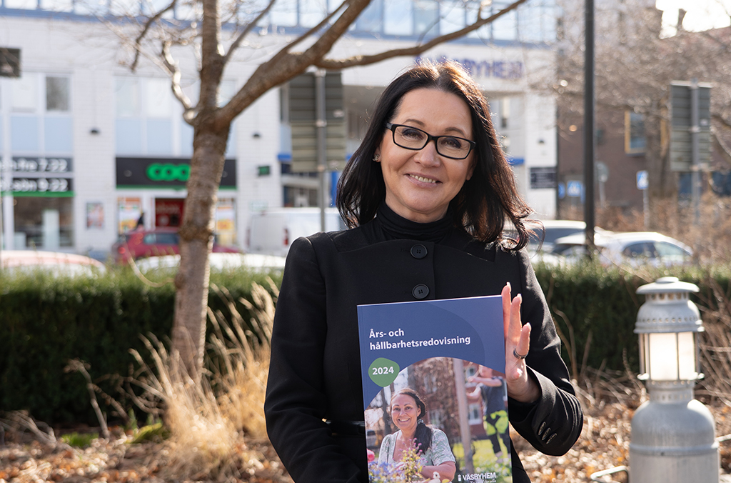 Kvinna i mörka kläder och glasögon stående utomhus med en års- och hållbarhetsrapport i händerna.