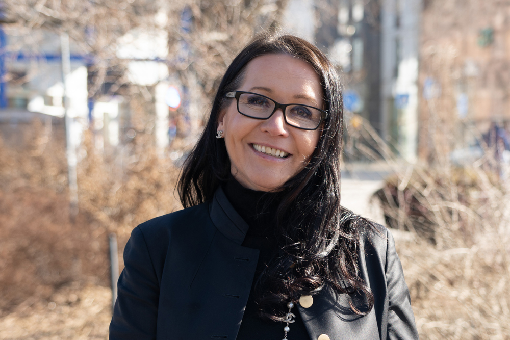 Leende kvinna med mörkt hår, svarta glasögon och svarta kläder i stadsmiljö.