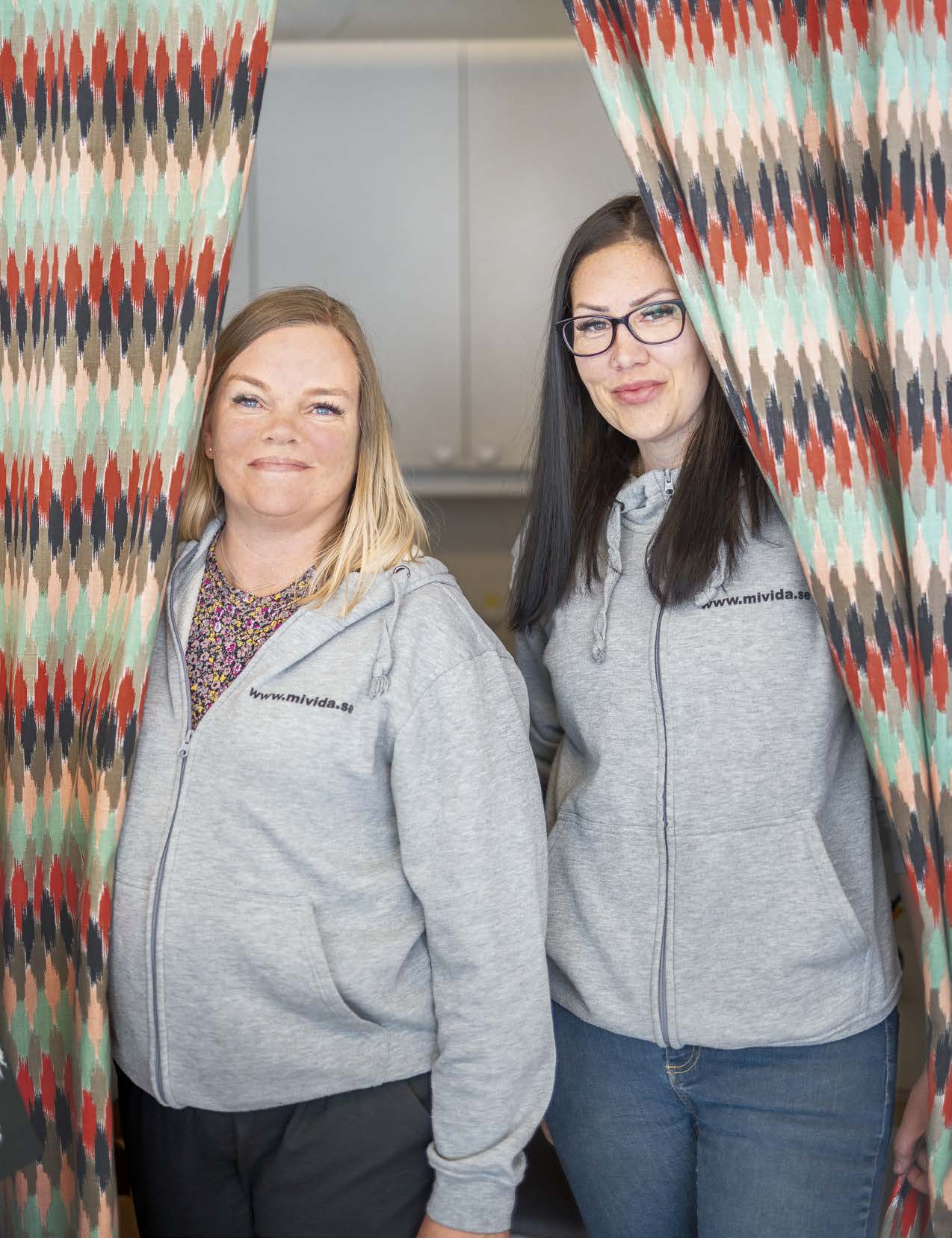 Två kvinnor står intill varandra mellan ett draperi i olika färger med köksluckor i bakgrunden