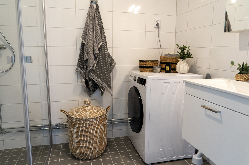 Badrum med vitt kakel och grått klinker, duschväggar i glas, tvättkorg med lock, grå handdukar, tvättmaskin och handfatsmöbel med låda. 