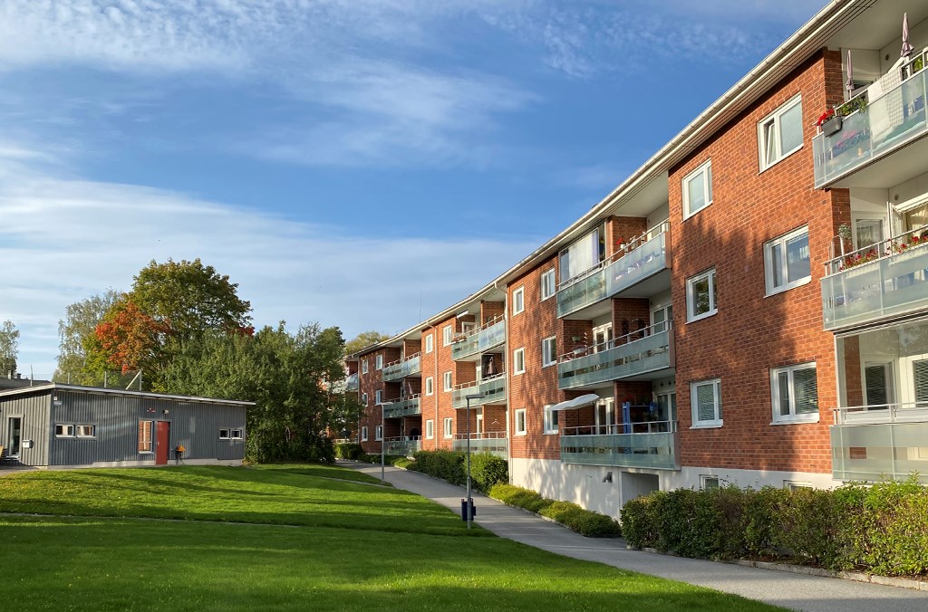 Trevånings flerbostadshus i tegel med balkongfronter i glas invid en gräsmatta och en tvättstuga.