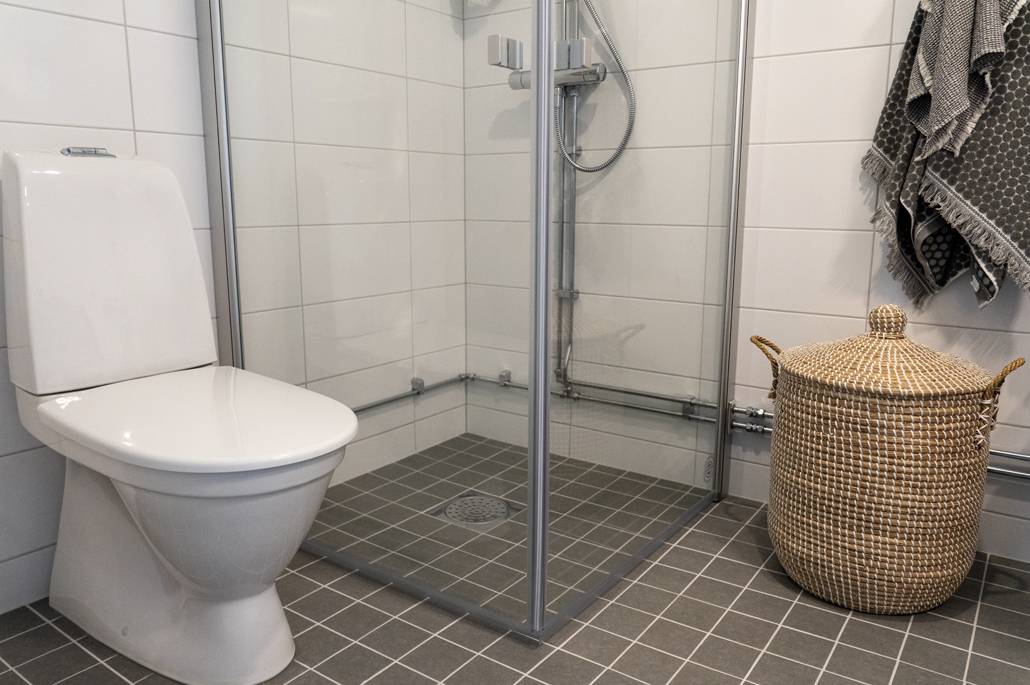 Badrum med vitt kakel, grått klinker, duschväggar i glas, toalettstol, tvättkorg i naturfärgat material och grå handdukar.