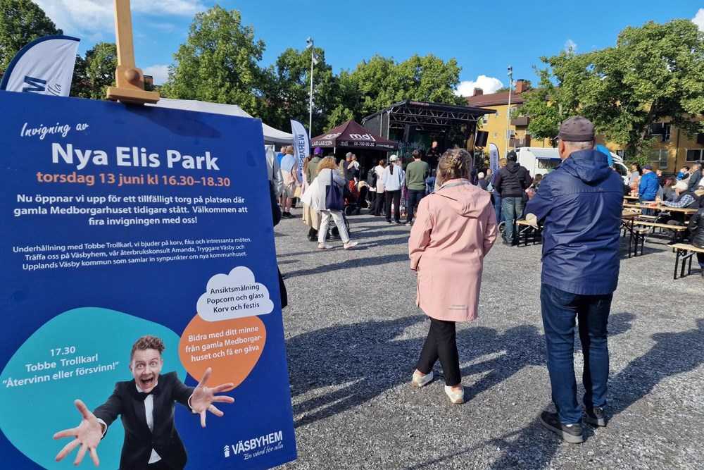 Skylt med texten Invigning av Nya Elis Park i förgrunden med människor stående framför en scen.