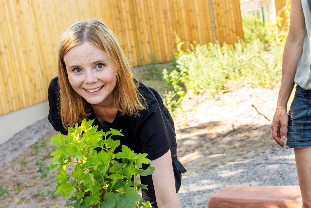 Kvinna i svart t-shirt på knä invid en grön planta med två personer stående intill.