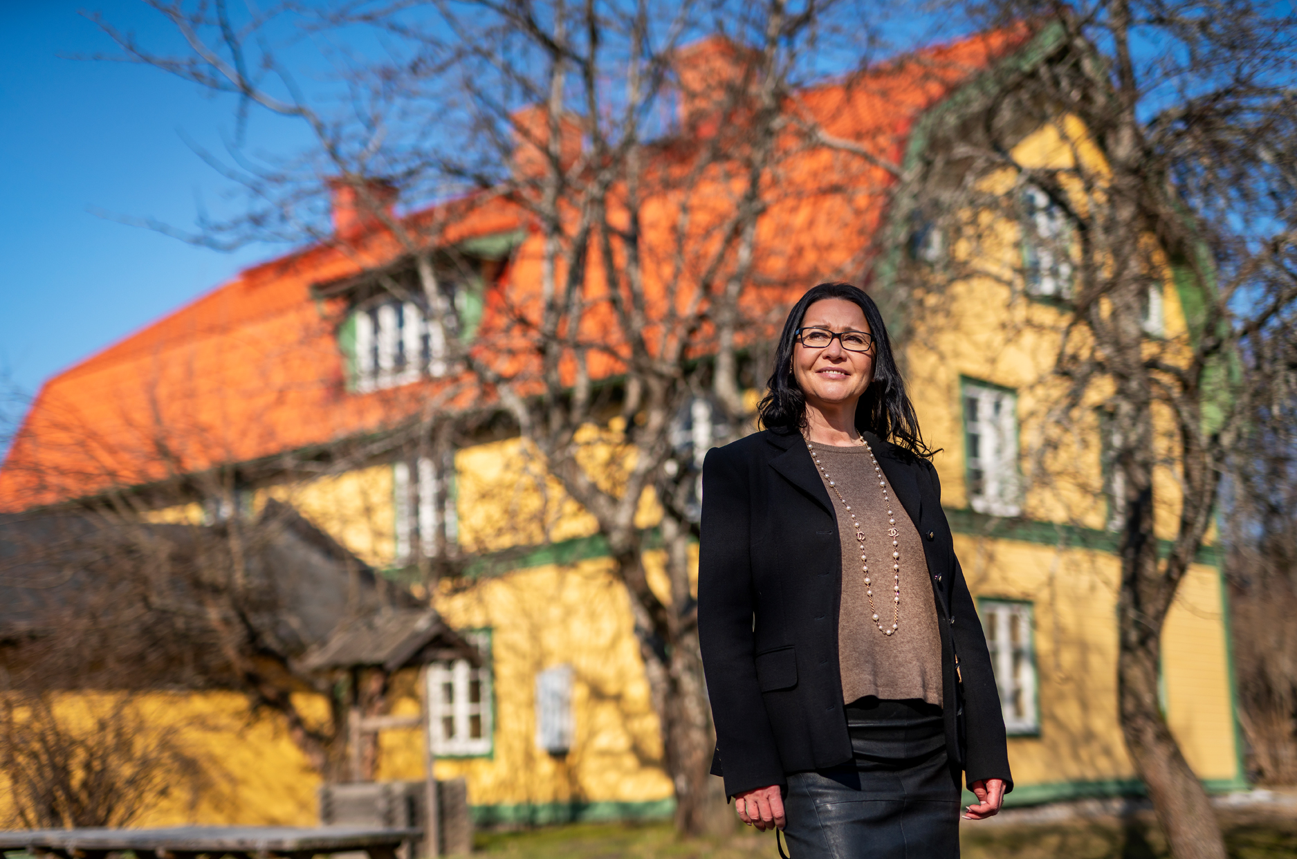Kvinna i svart kavaj står utomhus i solsken framför ett gult trähus.