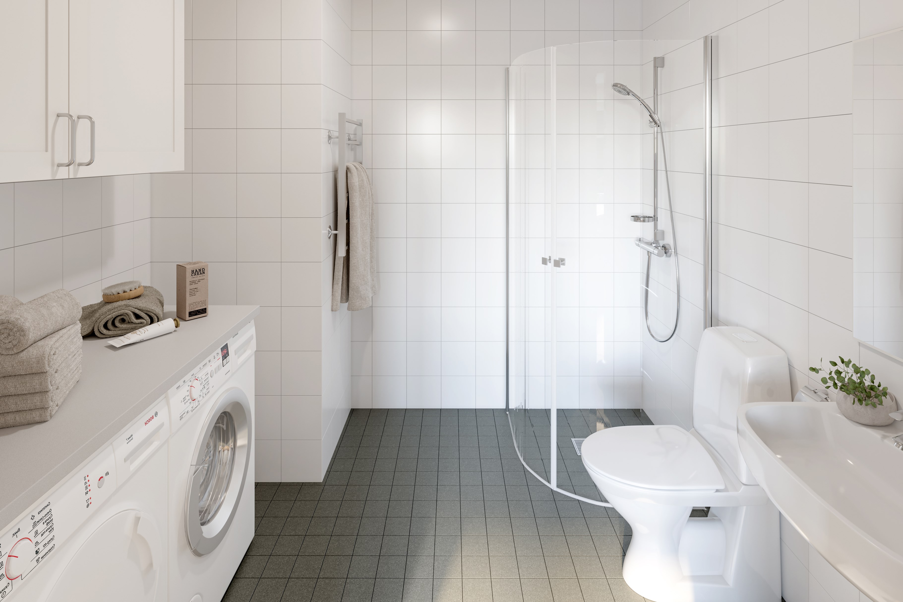 Badrum med grå klinker på golv, vitt kakel på väggar, duschväggar i glas, vit toalettstol och handfat, vita överskåp och bänkskiva med tvättmaskin och torktumlare under.