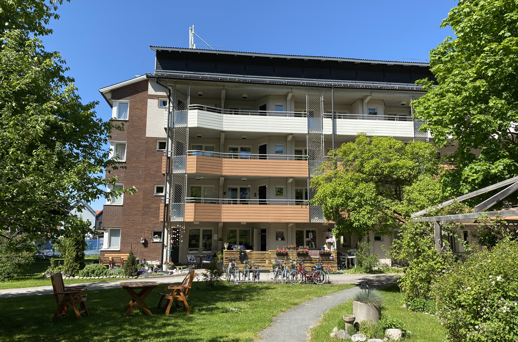 Grönskande gård med sittgrupp, cykelställ, pergola, träd och buskar framför ett flerbostadshus med loftgångar i brunt