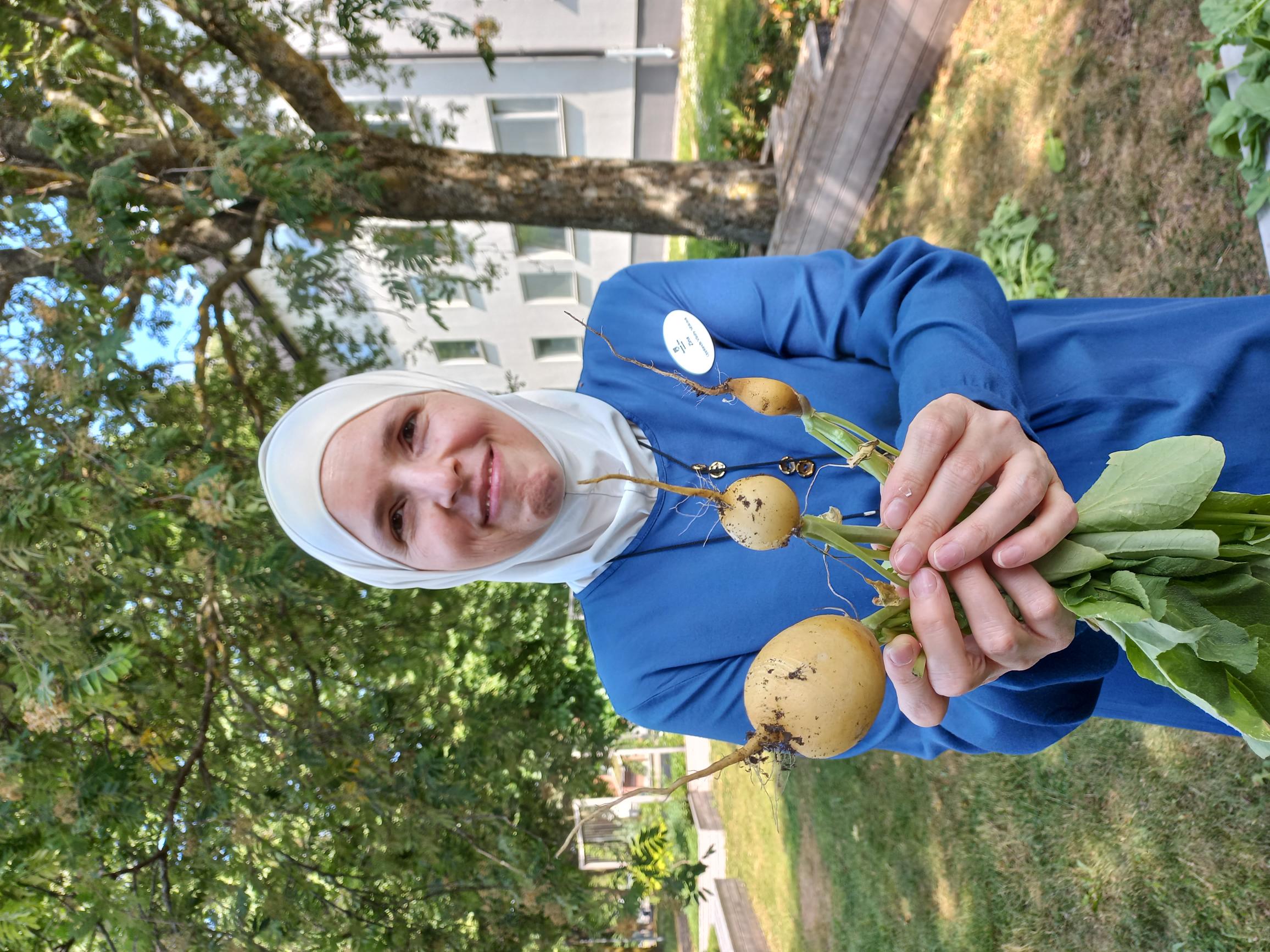 En leende kvinna stående framför en grönskande bakgrund håller upp tre vita rädisor framför sig. 