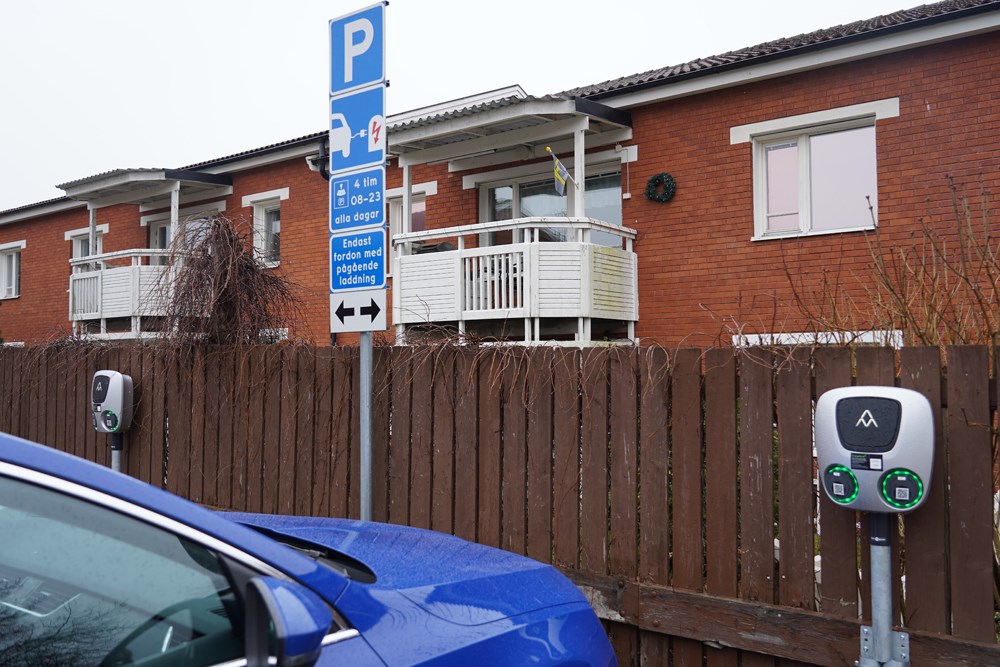 Blå personbil intill laddplats för elbil med brunt staket och bostadshus i tegel i bakgrunden.