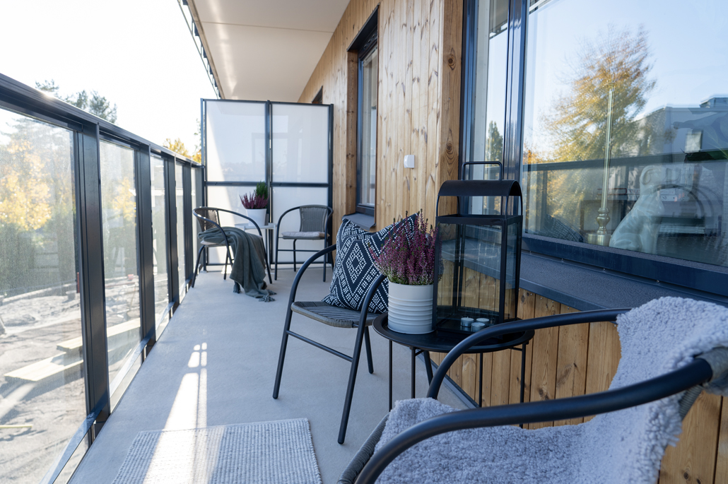 Insidan av en balkong med räcke i glas, stolar av svart stål kring litet svart bord med en blomkruka med lila ljung i.